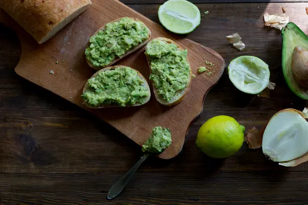 Preparación de aguacate guacamole en proceso todos los ingridientes sobre lima de mesa, cebolla, aguacate todo orgánico y fresco sobre fondo rústico de madera oscura con guacamole listo en una toma de ángulo superior de enfoque suave de pan de ciabatta horneado fresco — Foto de Stock