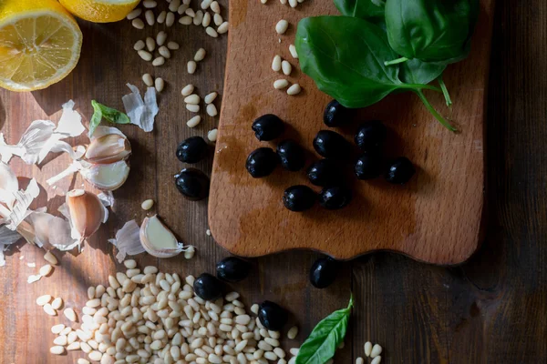 Italian pesto preparation vegan version without parmesan cheese with black olives instead all ingredients on table green basil, garlic, lemon, pine nuts, black olives all fresh and organic on dark wood rustic style table background soft focus — Stock Photo, Image