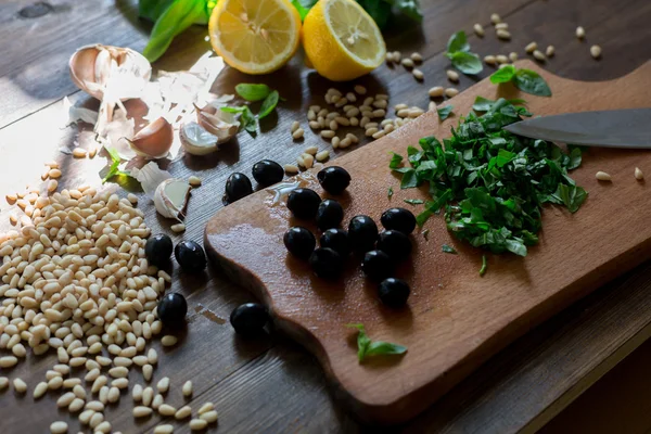 Italian pesto preparation vegan version without parmesan cheese with black olives instead all ingredients on table green basil, garlic, lemon, pine nuts, black olives all fresh and organic on dark wood rustic style table background soft focus — Stock Photo, Image