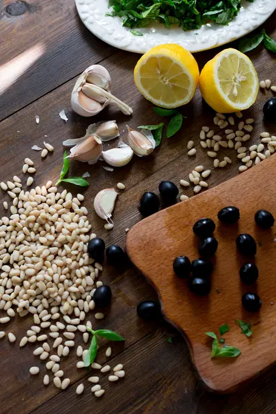 Italian pesto preparation vegan version without parmesan cheese with black olives instead all ingredients on table green basil, garlic, lemon, pine nuts, black olives all fresh and organic on dark wood rustic style table background soft focus — Stock Photo, Image