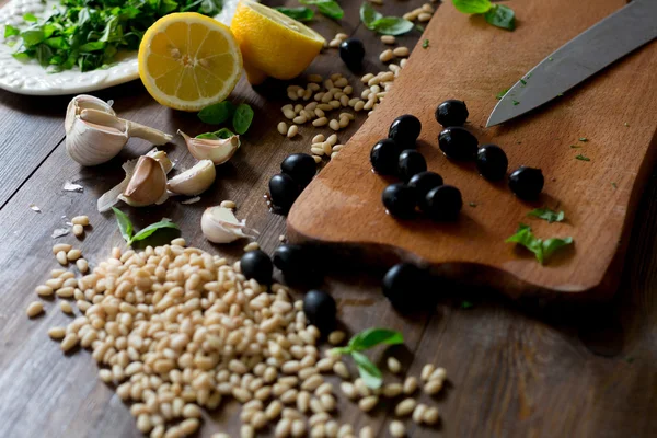 Italian pesto preparation vegan version without parmesan cheese with black olives instead all ingredients on table green basil, garlic, lemon, pine nuts, black olives all fresh and organic on dark wood rustic style table background soft focus — Stock Photo, Image