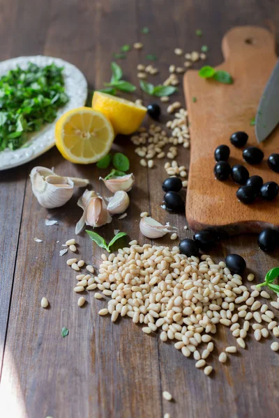 Italian pesto preparation vegan version without parmesan cheese with black olives instead all ingredients on table green basil, garlic, lemon, pine nuts, black olives all fresh and organic on dark wood rustic style table background soft focus — Stock Photo, Image