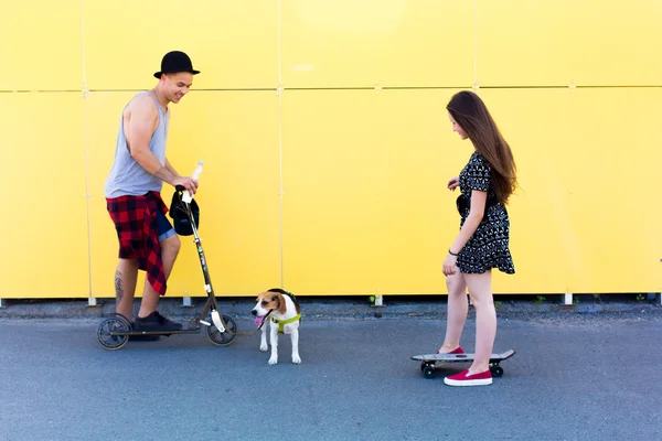 Fresco joven y hermosa rubia caucásica adolescente hipster skater chica con el pelo largo y hermoso chico hipster con su perro perrito beagle se divierten al aire libre y patinar con monopatín durante el increíble día de verano sobre un fondo amarillo — Foto de Stock