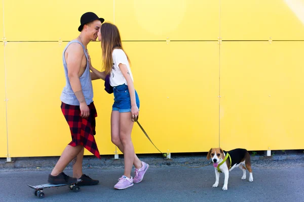 Cool young and beautiful caucasian blonde hipster girl wearing denim shorts with her handsome boyfriend on skate wearing a hat and beagle puppy dog posing smiling kissing having fun and skating outside as a loving perfect hipster couple — Stock Photo, Image