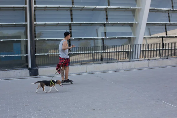 Cool young and handsome caucasian brunette hipster skater guy wearing a hat posing smiling and having fun outside while skating with his skateboard during amazing summer day in the city with his beagle pupppy dog — Stock Photo, Image