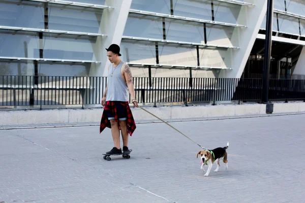 Cool mladý a pohledný kavkazské bruneta bederní bruslař chlap nosí klobouk pózuje s úsměvem a baví mimo při bruslení s jeho skateboard během úžasné letní den v městě se psem pupppy Bígl — Stock fotografie