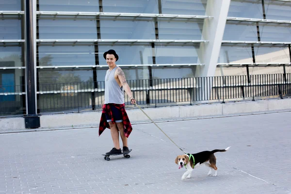 Cool young and handsome caucasian brunette hipster skater guy wearing a hat posing smiling and having fun outside while skating with his skateboard during amazing summer day in the city with his beagle pupppy dog — Stock Photo, Image