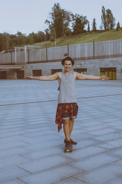 Fresco joven y guapo caucásico morena hipster skater chico usando un sombrero posando sonriendo y divertirse al aire libre mientras patina con su monopatín durante increíble día de verano en la ciudad —  Fotos de Stock