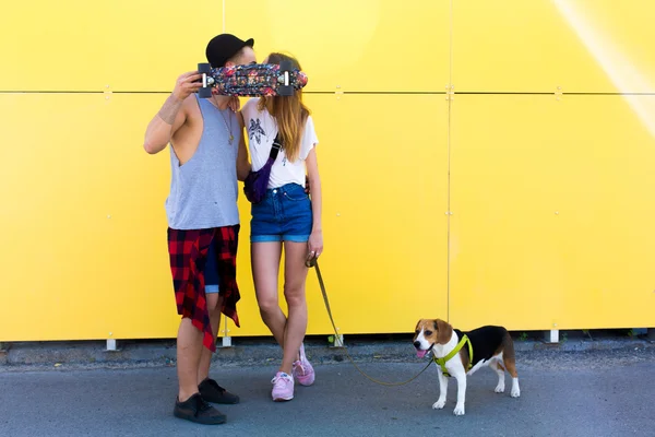 Fresco joven y hermosa chica hipster rubia caucásica usando pantalones cortos de mezclilla con su novio guapo en el patín con un sombrero y un perro perrito beagle posando besos sonrientes divertirse y patinar fuera como una pareja hipster perfecto amoroso — Foto de Stock