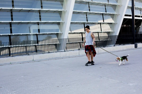 Cool young and handsome caucasian brunette hipster skater guy wearing a hat posing smiling and having fun outside while skating with his skateboard during amazing summer day in the city with his beagle puppy dog — Stock Photo, Image