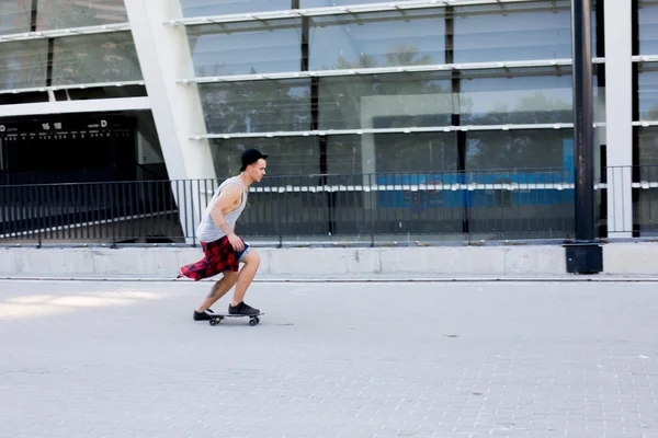 Cool mladý a pohledný kavkazské bruneta bederní bruslař chlap klobouk pózování s úsměvem a baví mimo při bruslení s jeho skateboard během úžasné letní den v městě — Stock fotografie