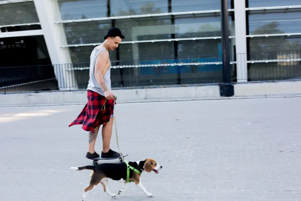 Cool young and handsome caucasian brunette hipster skater guy wearing a hat posing smiling and having fun outside while skating with his skateboard during amazing summer day in the city with his beagle puppy dog — Stock Photo, Image