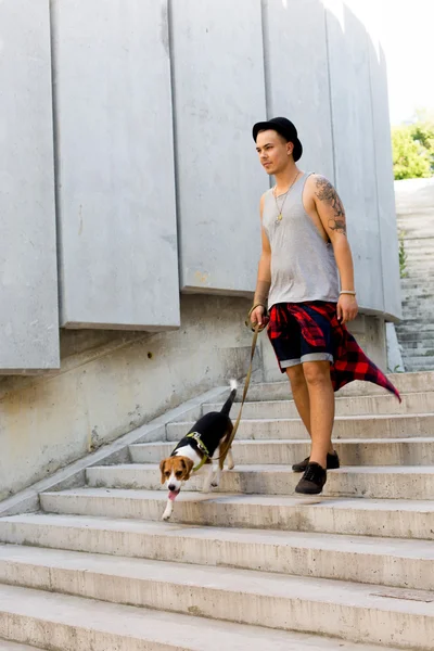 Cool young and handsome caucasian brunette hipster guy wearing a hat posing smiling and having fun outside during amazing summer day in the city with his beagle puppy dog — Stock Photo, Image