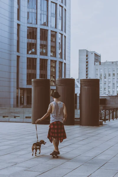 Fresco joven y guapo caucásico morena hipster skater chico usando un sombrero posando sonriente y divertirse al aire libre mientras patina con su monopatín durante increíble día de verano en la ciudad con su perro perrito beagle — Foto de Stock