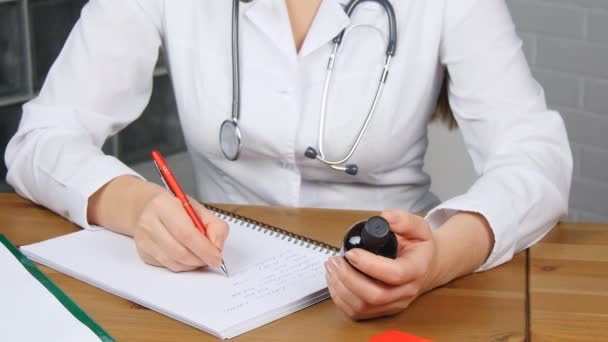 Hermosa joven doctora sentada frente a la mesa de trabajo sosteniendo frasco de tabletas y escribiendo receta en forma especial. Concepto médico y farmacéutico — Vídeos de Stock