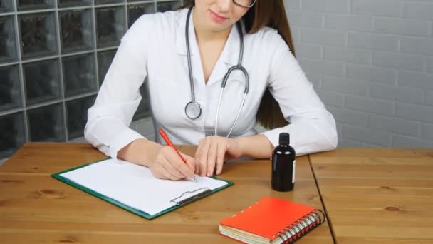 Vackra unga kvinnliga läkare sitter framför arbetsbord hålla burken med tabletter och skriva recept på särskild blankett. medicinsk och farmaceut koncept — Stockvideo