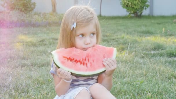 Милая маленькая девочка ест арбуз на траве в летнее время — стоковое видео