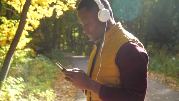 Концепция прослушивания музыки и подкастов. Улыбающийся молодой африканский американец слушает музыку в осеннем парке — стоковое видео