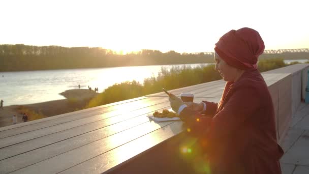 Молодая мусульманка, одетая в хиджаб, пьет кофе на свежем воздухе на фоне города. — стоковое видео
