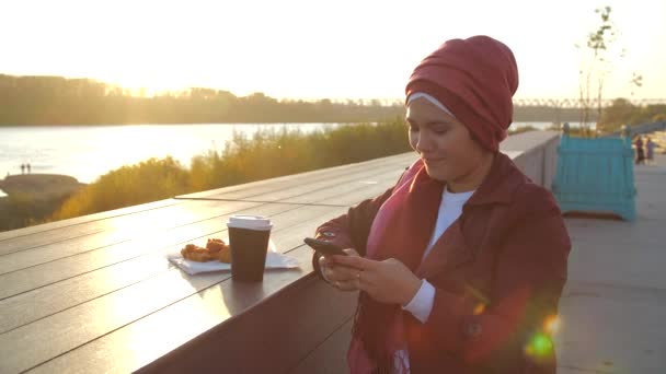 在城市背景下，年轻的穆斯林妇女戴着头巾，在户外喝咖啡休息 — 图库视频影像