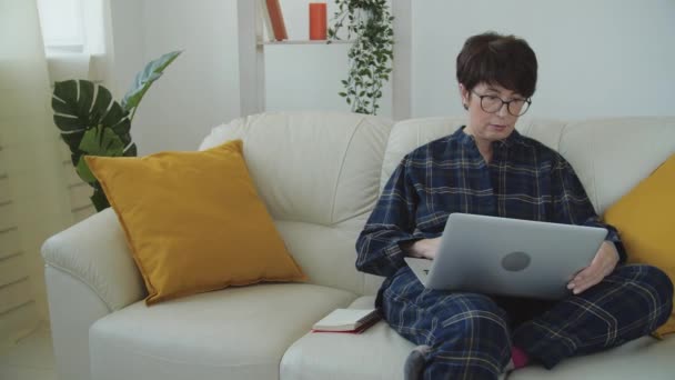 Conceito de trabalho remoto. Mulher madura de pijama trabalhando em casa com computador notebook — Vídeo de Stock