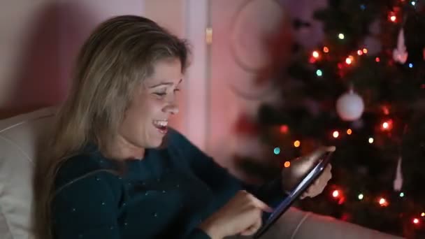 Jovem mulher sentada no sofá, sozinha, sobre a árvore de natal e usando tablet — Vídeo de Stock