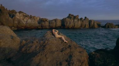 Genç beyaz çıplak kız deniz kıyısındaki kayalıklarda güneşleniyor. Altın bir günbatımının ılık ışınlarıyla okyanus boyunca süzülüyor. Kadınlar dinlenmeyi sever. Sabit çekim 