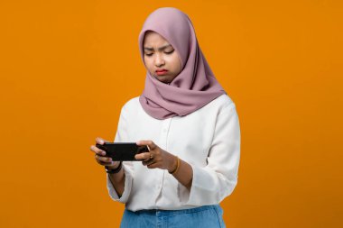 Üzgün genç Asyalı kadın sarı arka planda akıllı telefon kullanıyor.