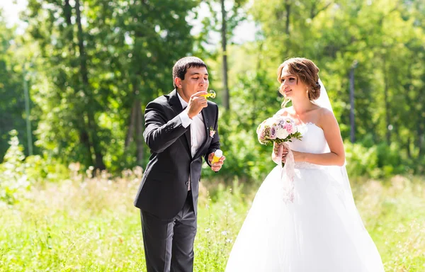 Elegante noivo elegante e noiva linda feliz se divertir com o ventilador de bolhas ao ar livre no parque — Fotografia de Stock