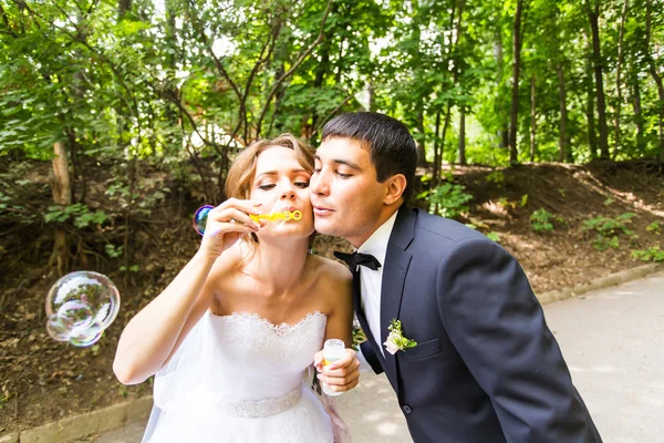 Pengantin pria dan pengantin wanita cantik yang elegan dan bahagia bersenang-senang dengan gelembung blower outdoors di taman — Stok Foto