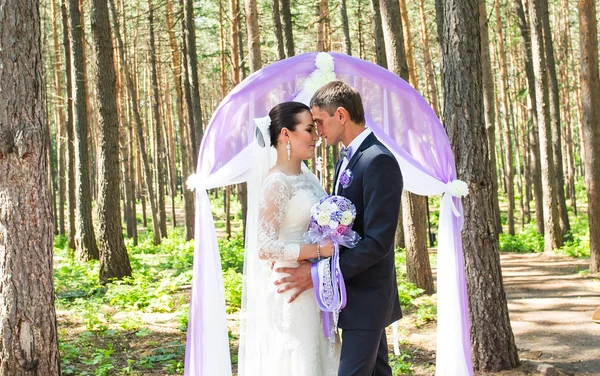 Nádherné stylové bohaté šťastné nevěsty a ženicha, stojící na svatební obřad v zelené zahradě poblíž fialové oblouk s květinami — Stock fotografie