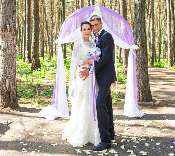 Csodálatos elegáns gazdag boldog menyasszony és a vőlegény állt egy esküvő, a zöld kert közelében lila virágok ív — Stock Fotó