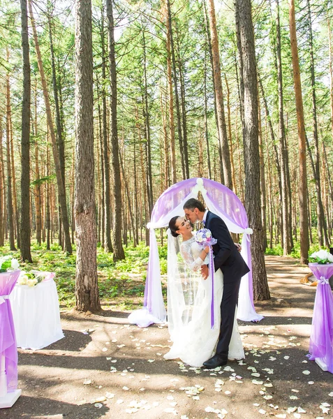 Maravilhosa noiva feliz rico elegante e noivo de pé em uma cerimônia de casamento no jardim verde perto do arco roxo com flores — Fotografia de Stock
