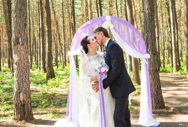 Csodálatos elegáns gazdag boldog menyasszony és a vőlegény állt egy esküvő, a zöld kert közelében lila virágok ív — Stock Fotó