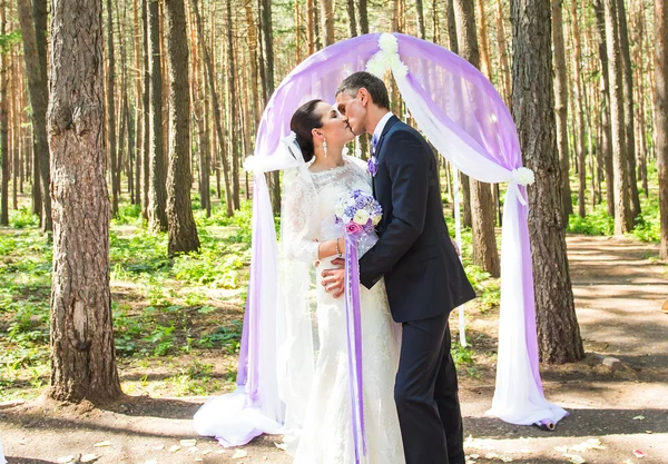 Underbar snygg rika lyckliga brudparet kysser på ett bröllop i grönskande trädgård nära lila båge med blommor — Stockfoto