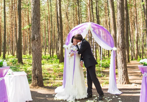 Maravilloso elegante rica novia feliz y novio besándose en una ceremonia de boda en jardín verde cerca de arco púrpura con flores —  Fotos de Stock