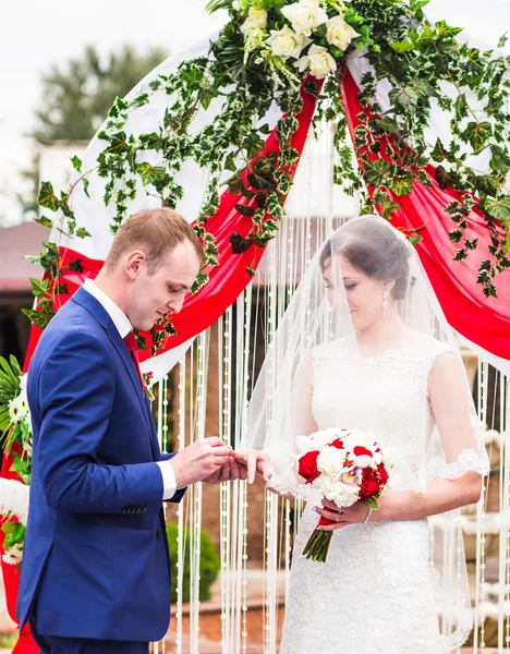 Casal em anéis de troca de trajes de casamento com arco no fundo — Fotografia de Stock