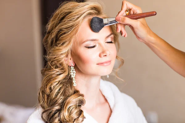 Artista de maquiagem de casamento fazendo uma maquiagem para a noiva. Menina modelo sexy bonita dentro de casa. Mulher loira beleza com cabelo encaracolado. Close-up mãos perto do rosto — Fotografia de Stock