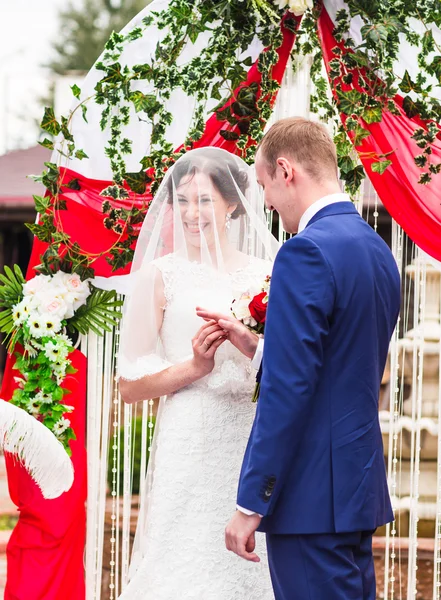 Couple in wedding attire exchange rings   with arch on background — Stock Photo, Image