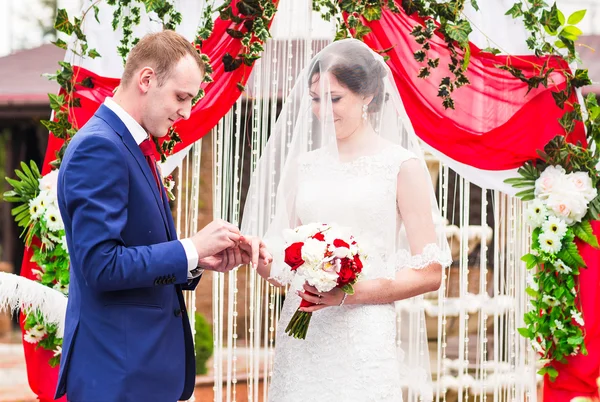 Casal em anéis de troca de trajes de casamento com arco no fundo — Fotografia de Stock