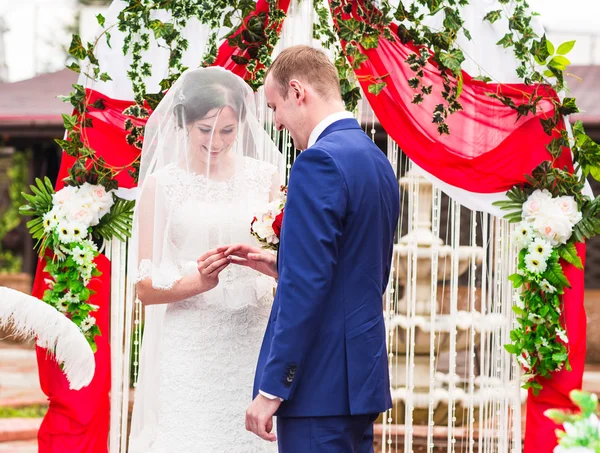 Casal em anéis de troca de trajes de casamento com arco no fundo — Fotografia de Stock