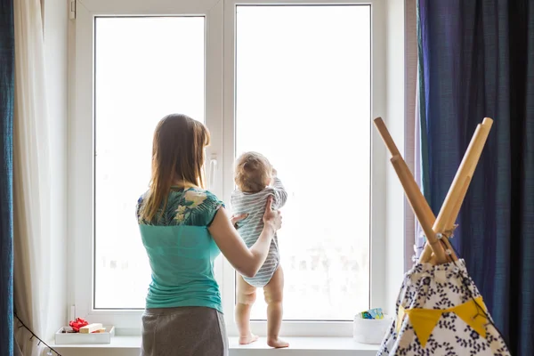 Explorer le monde ensemble. Vue latérale de la belle jeune femme tenant son bébé garçon tout en se tenant au rebord de la fenêtre à la maison — Photo