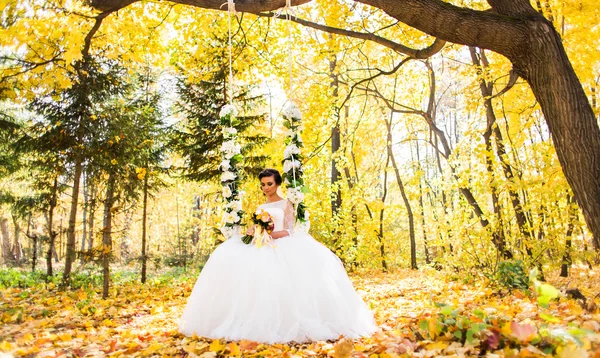 Prachtige bruid zit op de schommel in een herfst park — Stockfoto