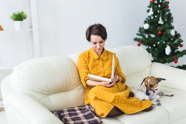A nő könyvet olvas a karácsonyfa előtt Jack Russell terrierrel. Karácsony, ünnepek és kisállat. — Stock Fotó