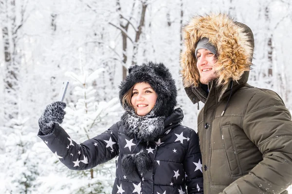 Sezon, aşk, teknoloji ve eğlence konsepti. Mutlu çift kış arkaplanı boyunca akıllı telefonla selfie çekiyor. — Stok fotoğraf