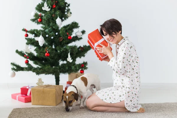 Glückliche Frau mit Hund öffnet Weihnachtsgeschenke. Weihnachtsbaum mit Geschenken darunter. Geschmücktes Wohnzimmer — Stockfoto