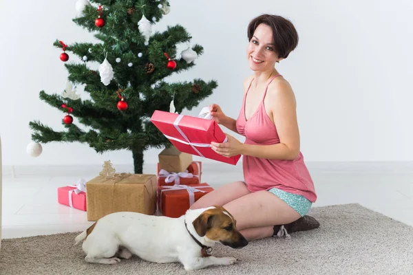 Ευτυχισμένη γυναίκα με σκύλο να ανοίγει χριστουγεννιάτικα δώρα. Χριστουγεννιάτικο δέντρο με δώρα από κάτω. Διακοσμημένο σαλόνι — Φωτογραφία Αρχείου