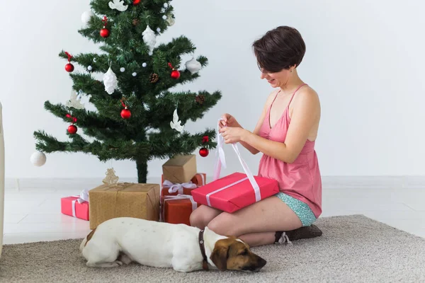 Ευτυχισμένη γυναίκα με σκύλο να ανοίγει χριστουγεννιάτικα δώρα. Χριστουγεννιάτικο δέντρο με δώρα από κάτω. Διακοσμημένο σαλόνι — Φωτογραφία Αρχείου
