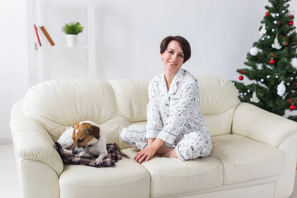 Ευτυχισμένη νεαρή γυναίκα που φοράει πιτζάμα με υπέροχο σκύλο στο σαλόνι με χριστουγεννιάτικο δέντρο. Έννοια διακοπών. — Φωτογραφία Αρχείου