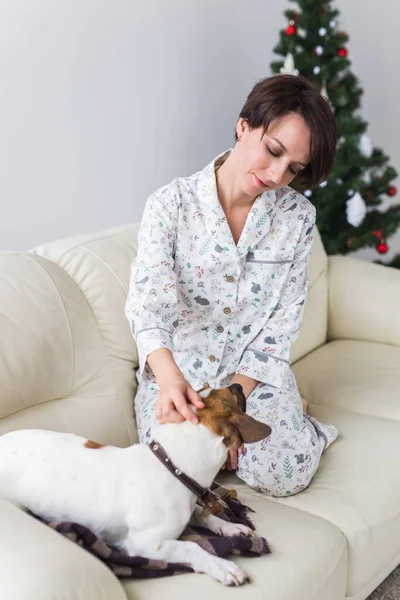 Joyeux jeune femme en pyjama avec beau chien dans le salon avec arbre de Noël. Concept de vacances. — Photo